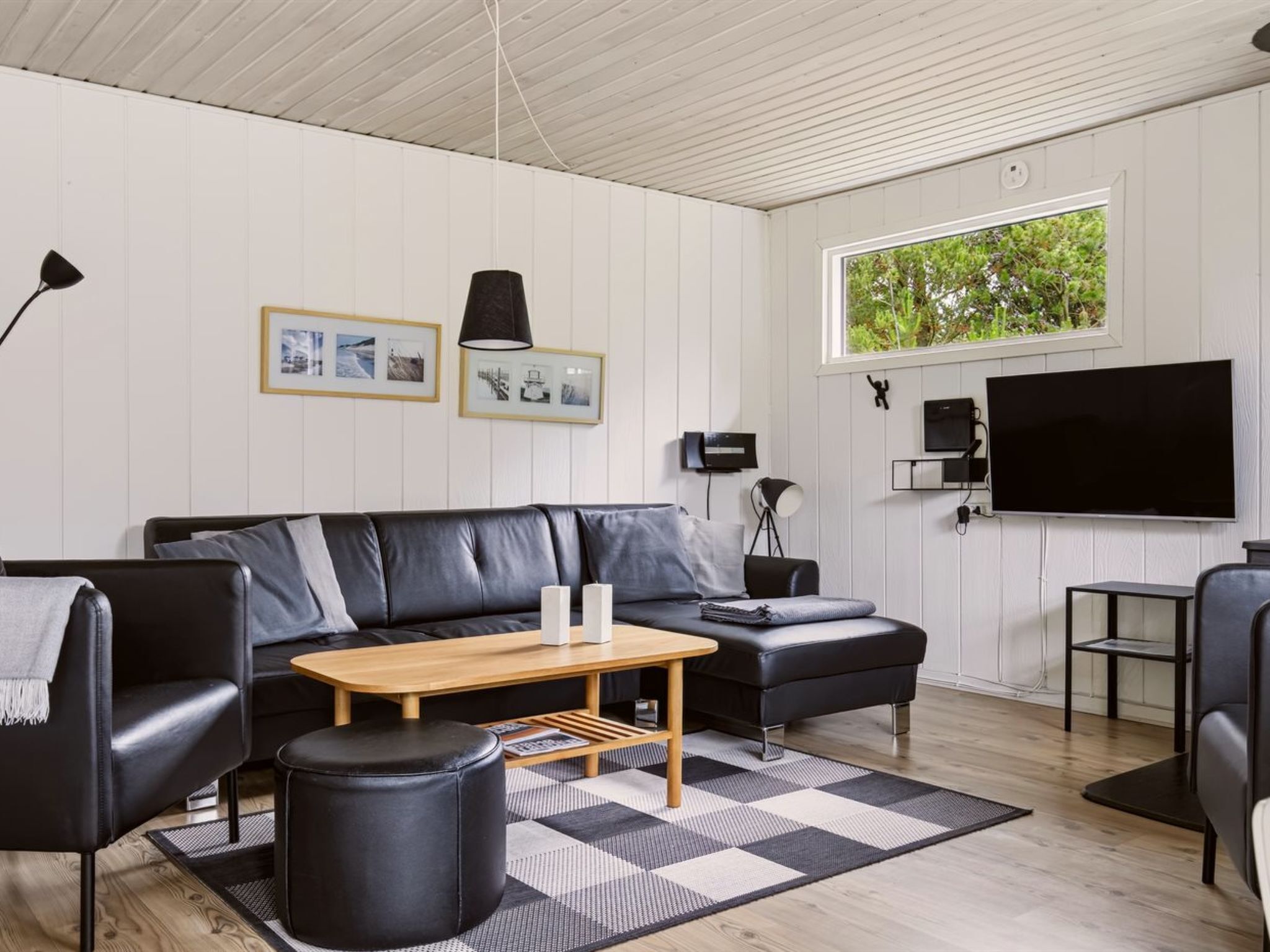 Een moderne woonkamer met een zwarte leren bank, een houten salontafel, een zwart poefje en een grote televisie aan de muur.