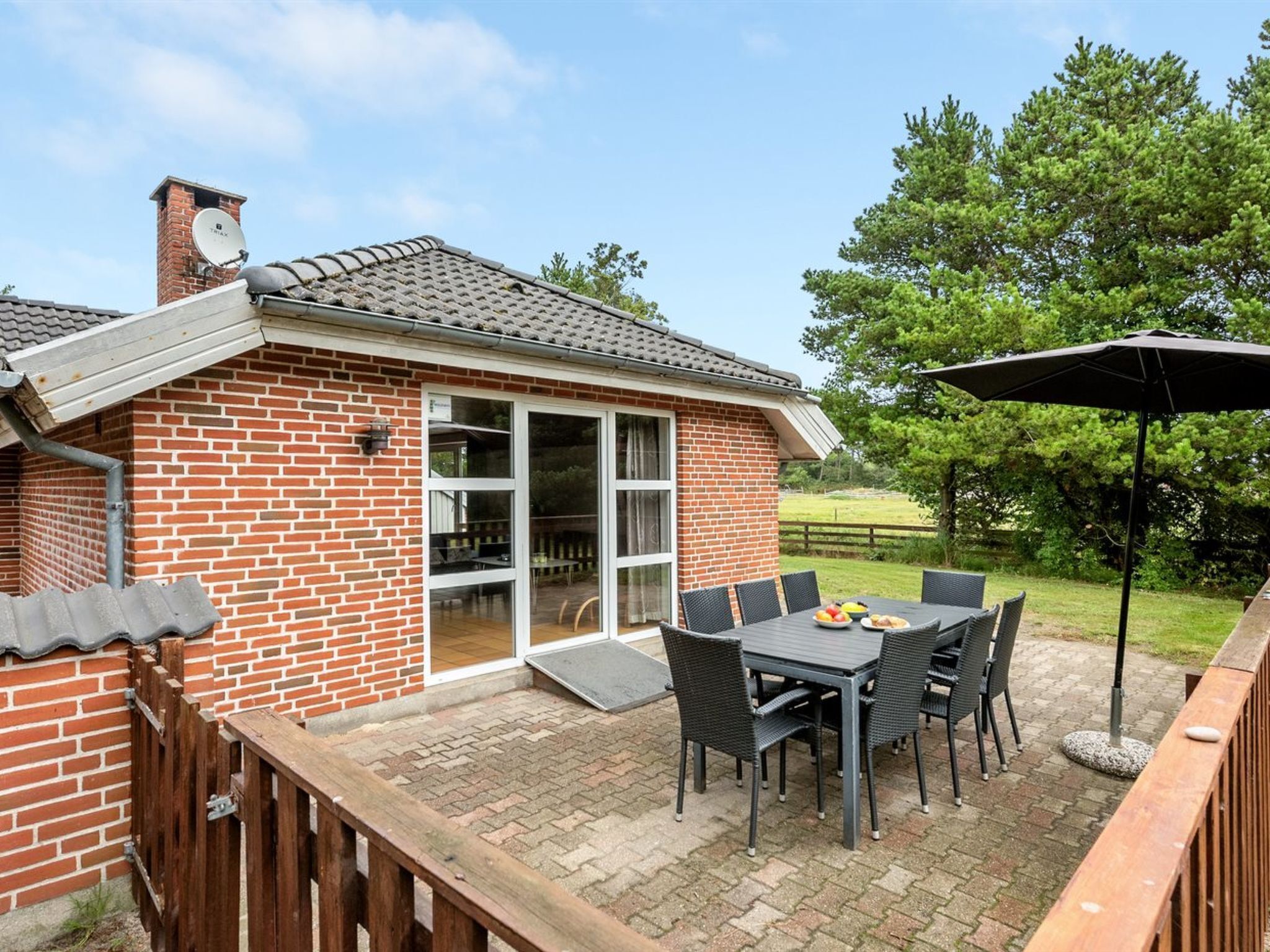 Een bakstenen huis met een patio, inclusief een tafel en stoelen, een houten terras en uitzicht op een groen landschap.