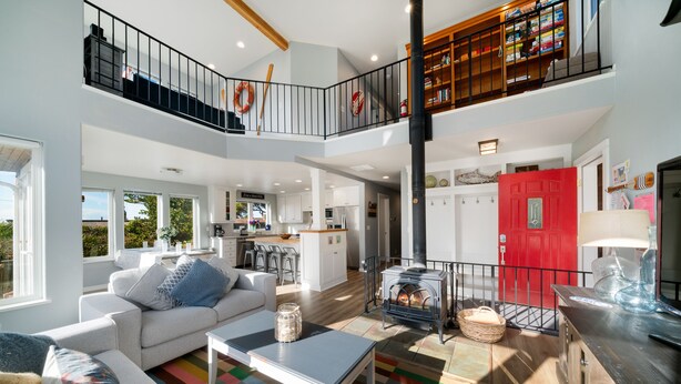 A spacious living area with a sofa, coffee table, and a colorful rug. A kitchen is visible in the background, and a red door is prominent. - The Cachalot MCA# 50330- Oceanview, 1/4 block to beach, Dogs OK, Game room (Manzanita)