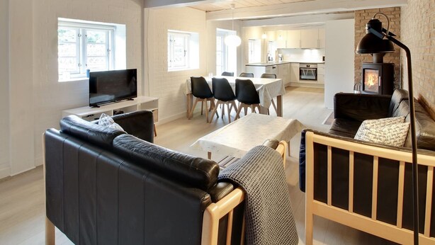 Una sala moderna con un sofá de cuero negro, un banco de madera y un televisor. Hay una zona de comedor con sillas y una mesa, y una cocina con estufa.