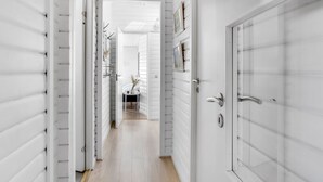 A narrow hallway with white paneled walls and a wooden floor, leading to a room with a small table and a chair.
