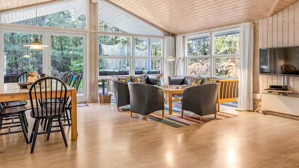 Een ruime kamer met een houten plafond, grote ramen, een eethoek met stoelen en een tafel, en een televisie aan de muur.