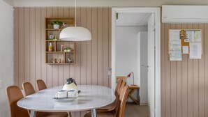 Een eethoek met een ronde tafel, bruine stoelen, een witte hanglamp, een houten plank met planten en een prikbord met papieren.