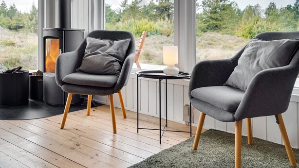 Een moderne kamer met twee grijze fauteuils, een kleine tafel met een lamp en een houtkachel.