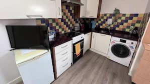 A compact kitchen with white cabinets, a black countertop, a stove, a washing machine, and a tiled backsplash. - Inviting 1-bed Apartment in Ilford (Ilford)