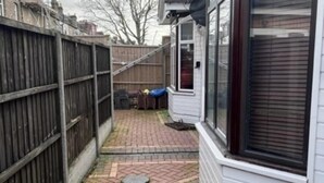 A residential backyard with a brick patio, a metal fence, and a window with blinds. - Inviting 1-bed Apartment in Ilford (Ilford)
