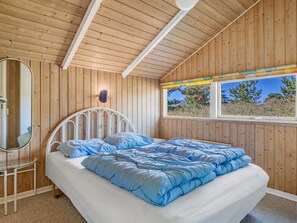 Camera da letto con soffitto in legno, un letto con biancheria azzurra, un comodino, uno specchio e una finestra con vista sugli alberi.