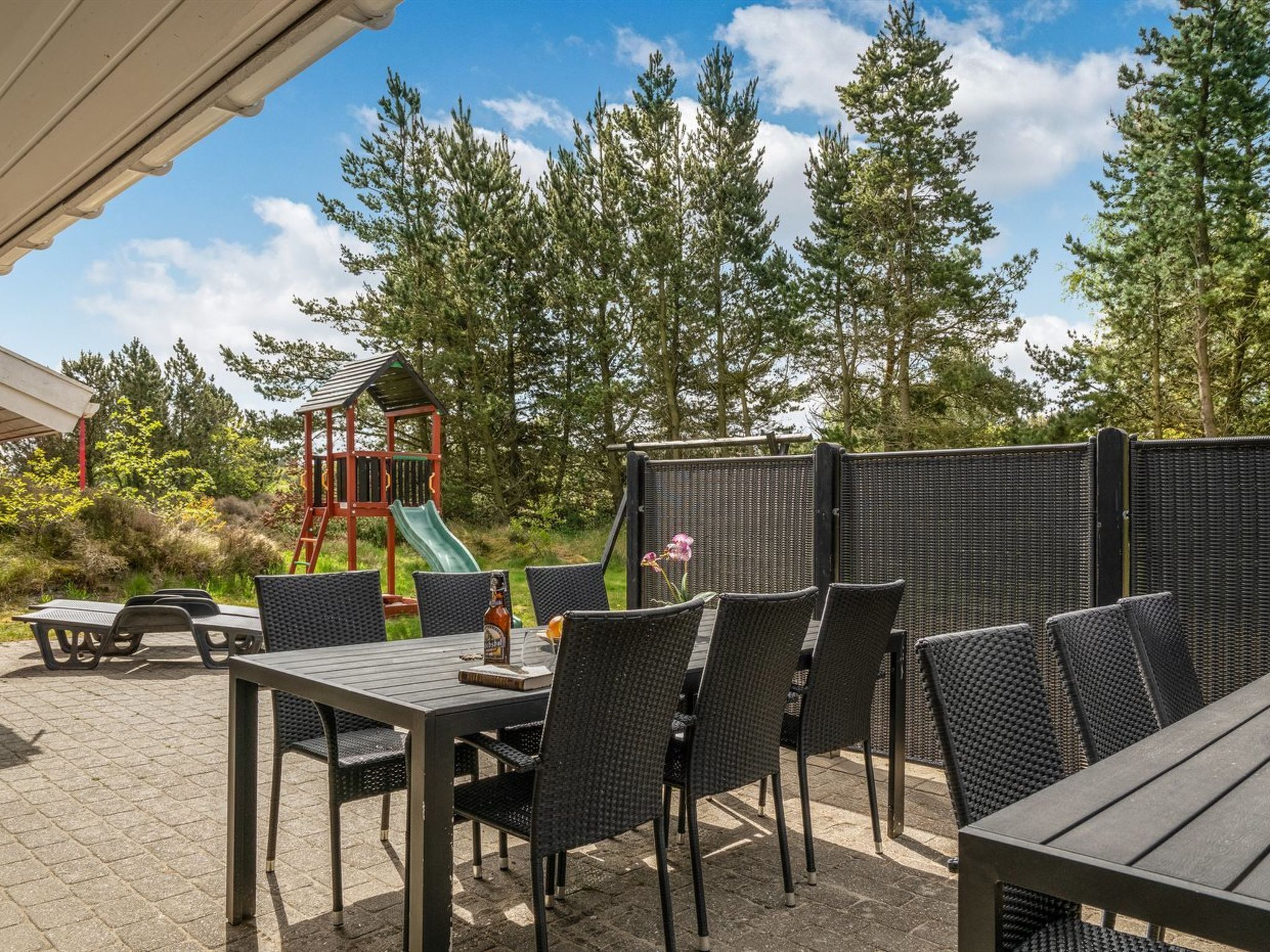 An outdoor patio with tables and chairs, a children's play structure, and a tall fence.