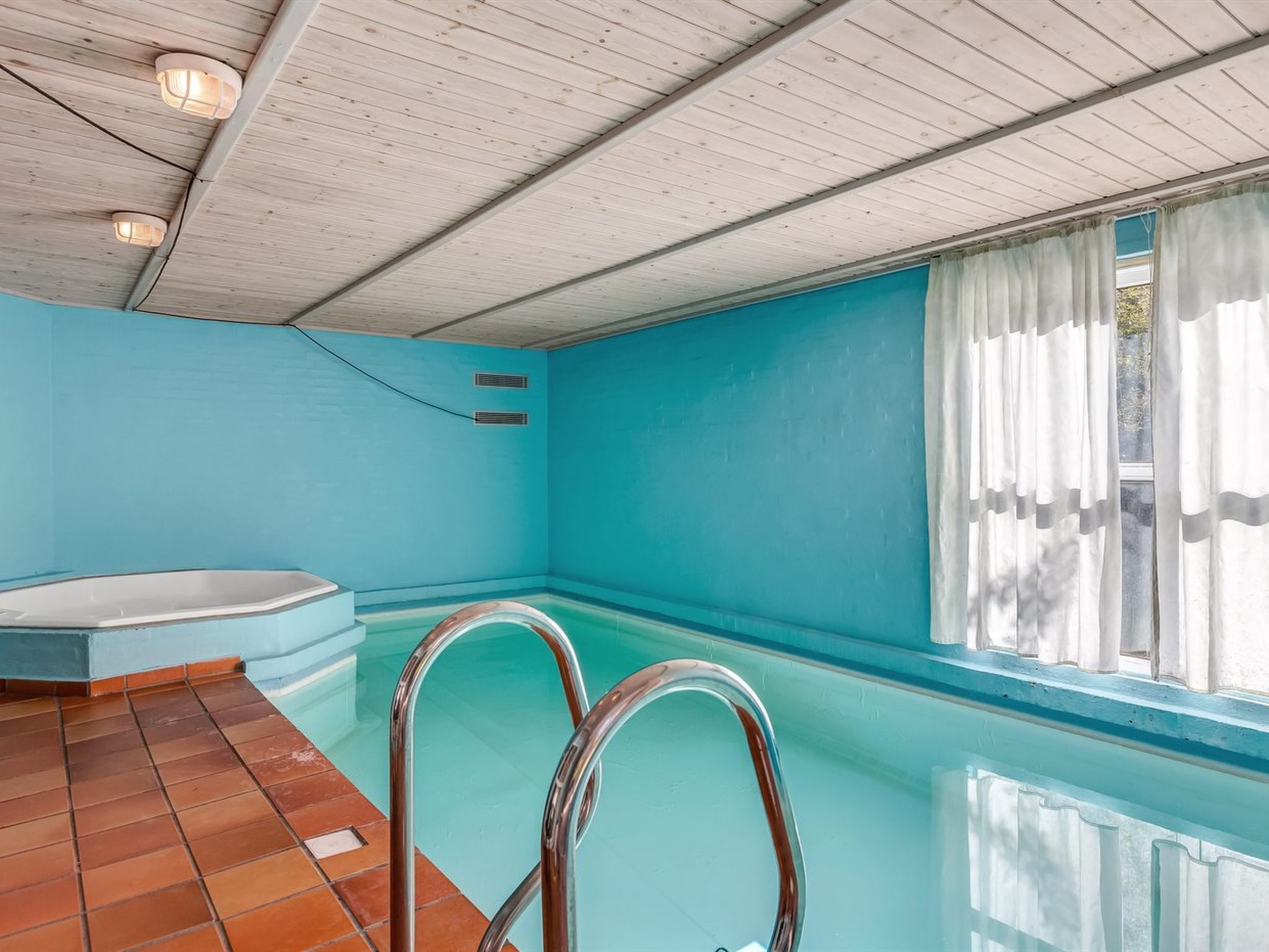 A swimming pool with clear water, a wooden ceiling, and a window with white curtains.