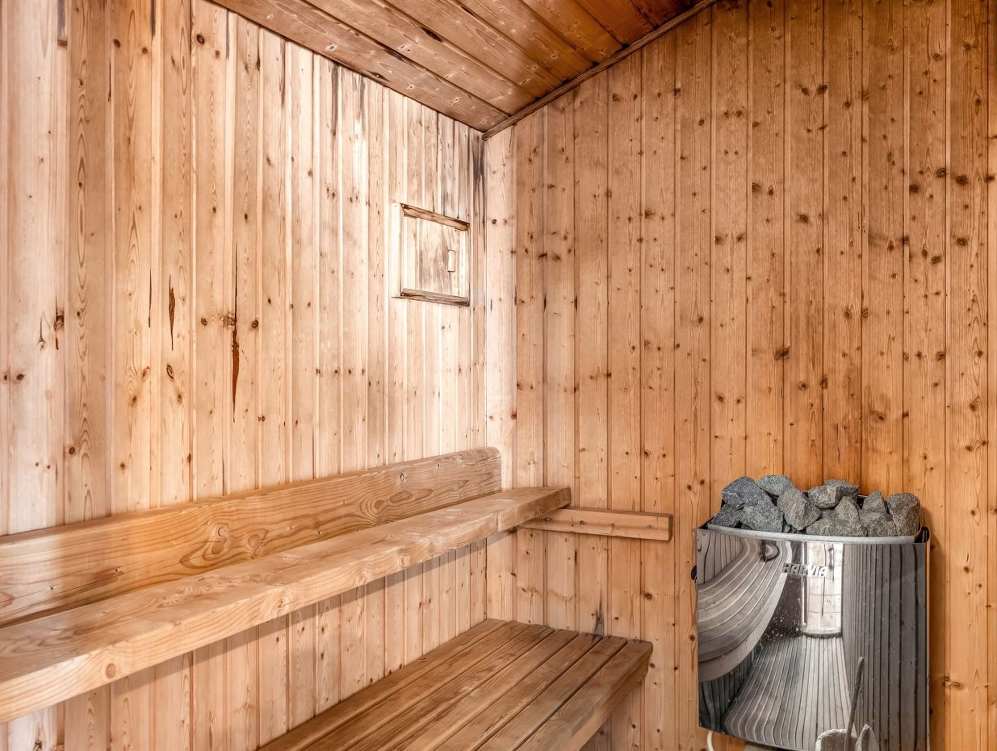 A sauna with wooden walls and benches, a metal container with stones, and a wooden shelf.