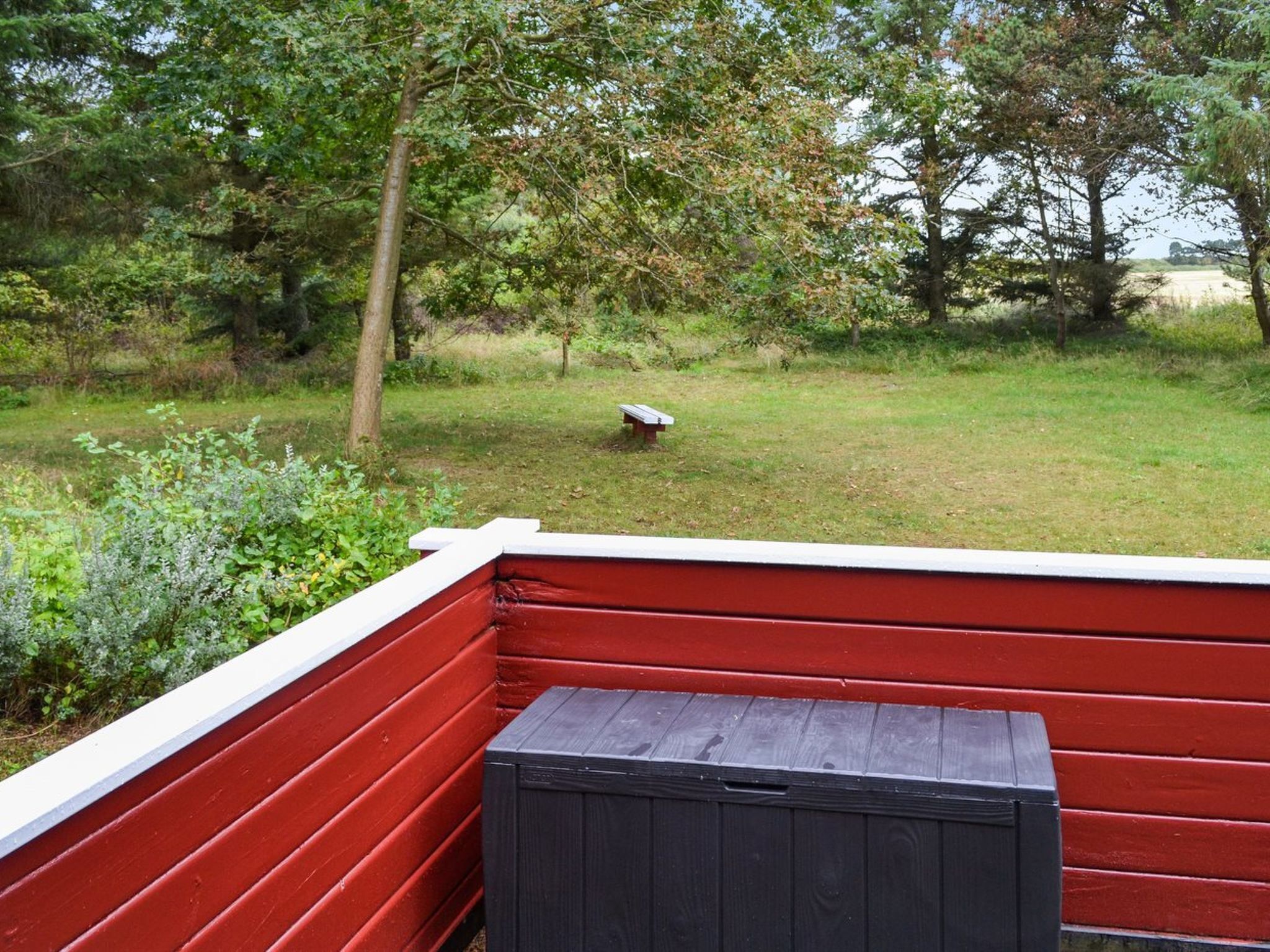 Eine rote Terrasse mit einer schwarzen Mülltonne, umgeben von einer grünen Fläche mit Bäumen und einer Bank in der Ferne.