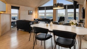 Ein modernes Interieur mit Holzboden, einem Essbereich mit weißem Tisch und schwarzen Stühlen, einer schwarzen Ledersofa und einem großen Fenster mit Blick ins Freie.
