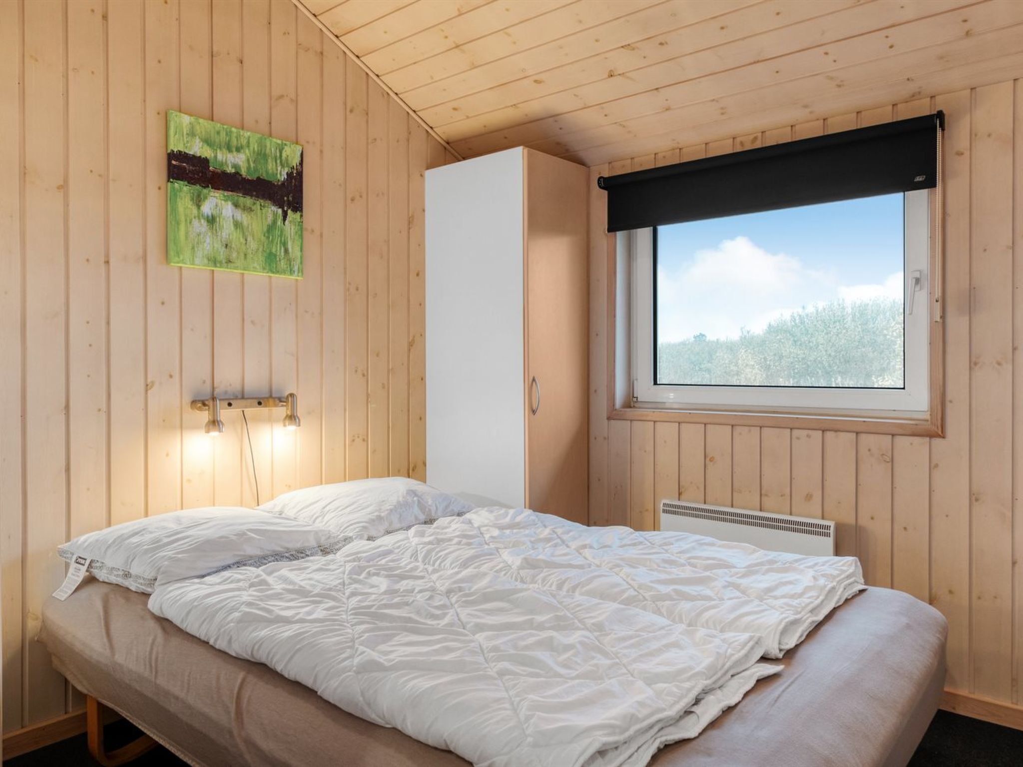 Une chambre à coucher avec un lit, une fenêtre donnant sur des arbres, une lampe fixée au mur et un tableau.