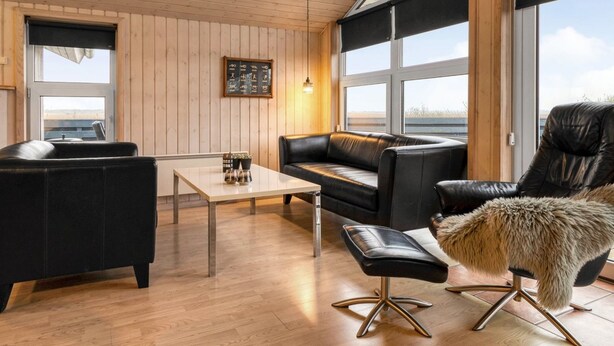 Ein modernes Wohnzimmer mit Holzbalkendecke, einem schwarzen Ledersofa, einem Ledersessel, einem Glastisch und einem großen Fenster mit Blick.