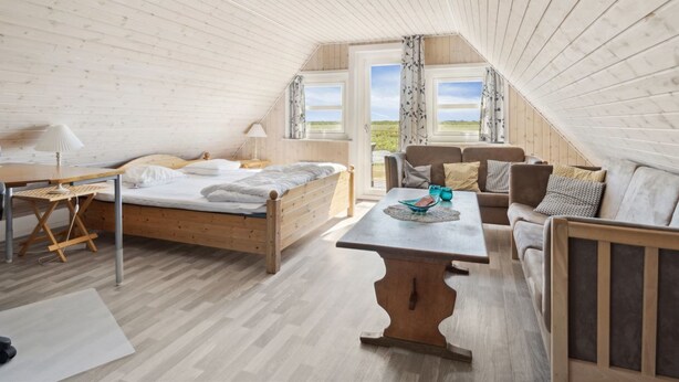 A cozy attic bedroom with a wooden ceiling, a bed, a sofa, a table, and a view of the outdoors.