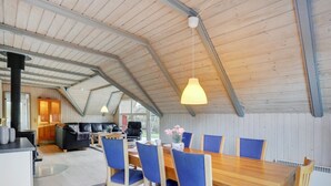 A spacious dining area with a wooden table and blue chairs, a kitchen in the background, and a high ceiling with exposed beams.