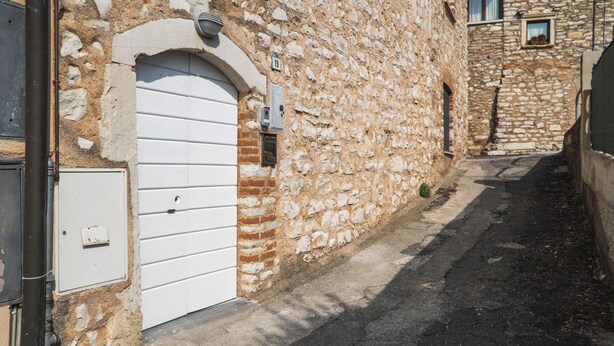 Rua estreita com edifícios de pedra, uma porta branca e uma pequena janela com uma planta.
