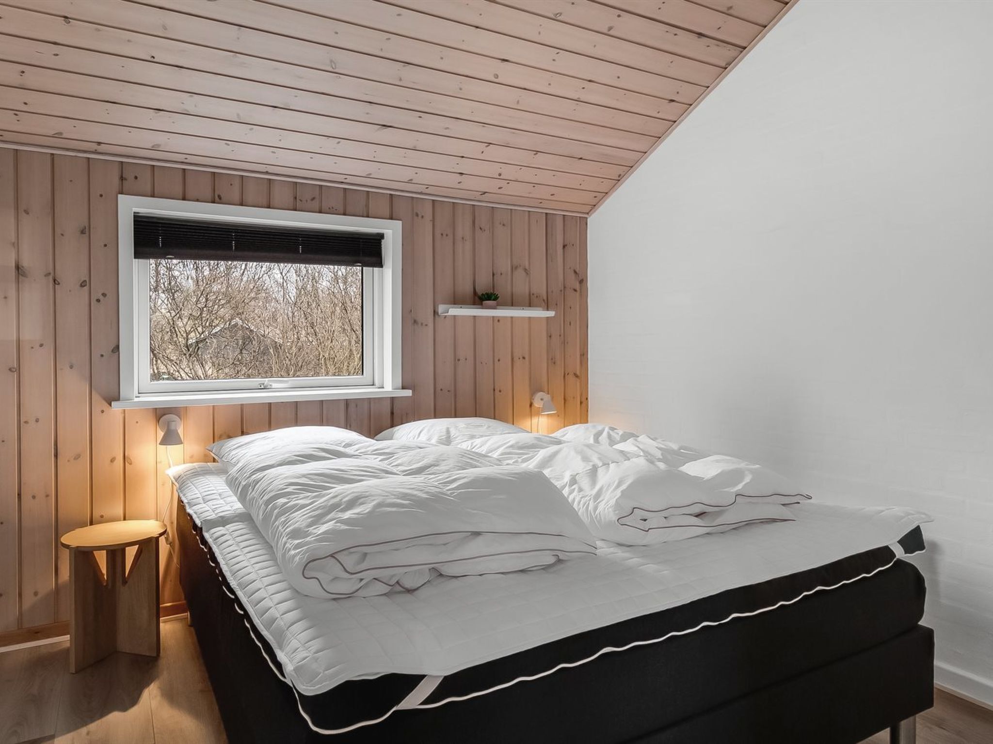 A bedroom with a wooden ceiling, a window with blinds, a bedside table, and a bed with white bedding.