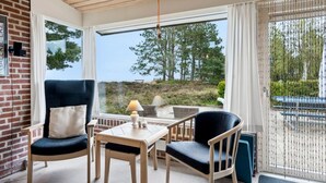 Una habitación con techo de madera, pared de ladrillo, un ventanal con vistas a árboles y una playa, una mesa con sillas y una lámpara colgante.