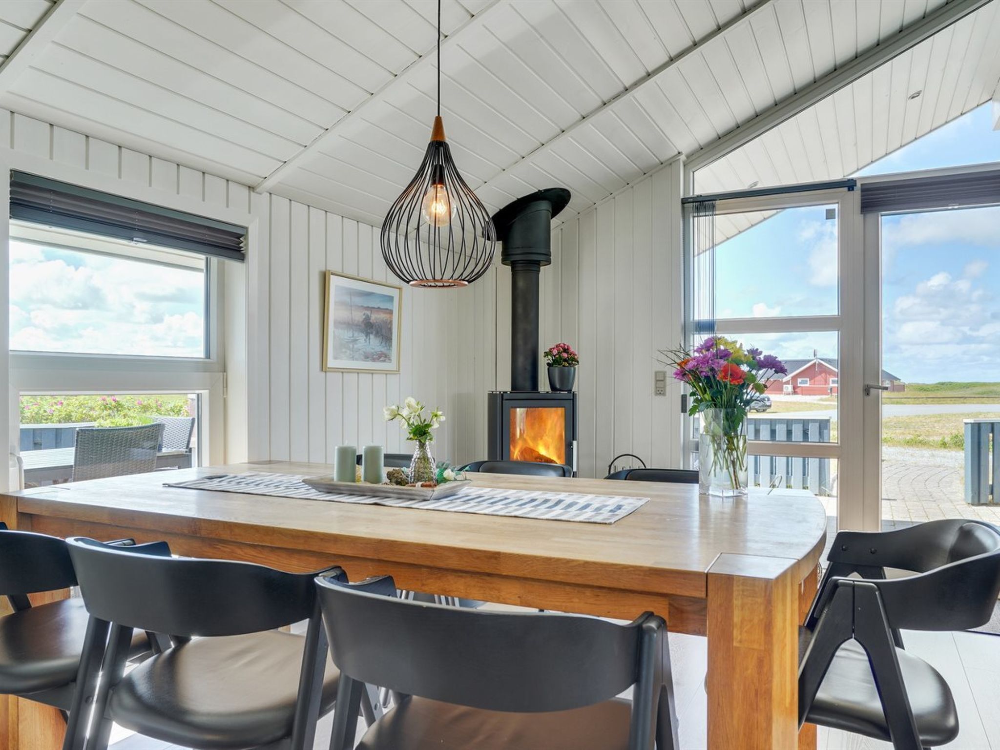 Un comedor moderno con una mesa de madera, sillas negras, una lámpara colgante, una chimenea y vistas al exterior.