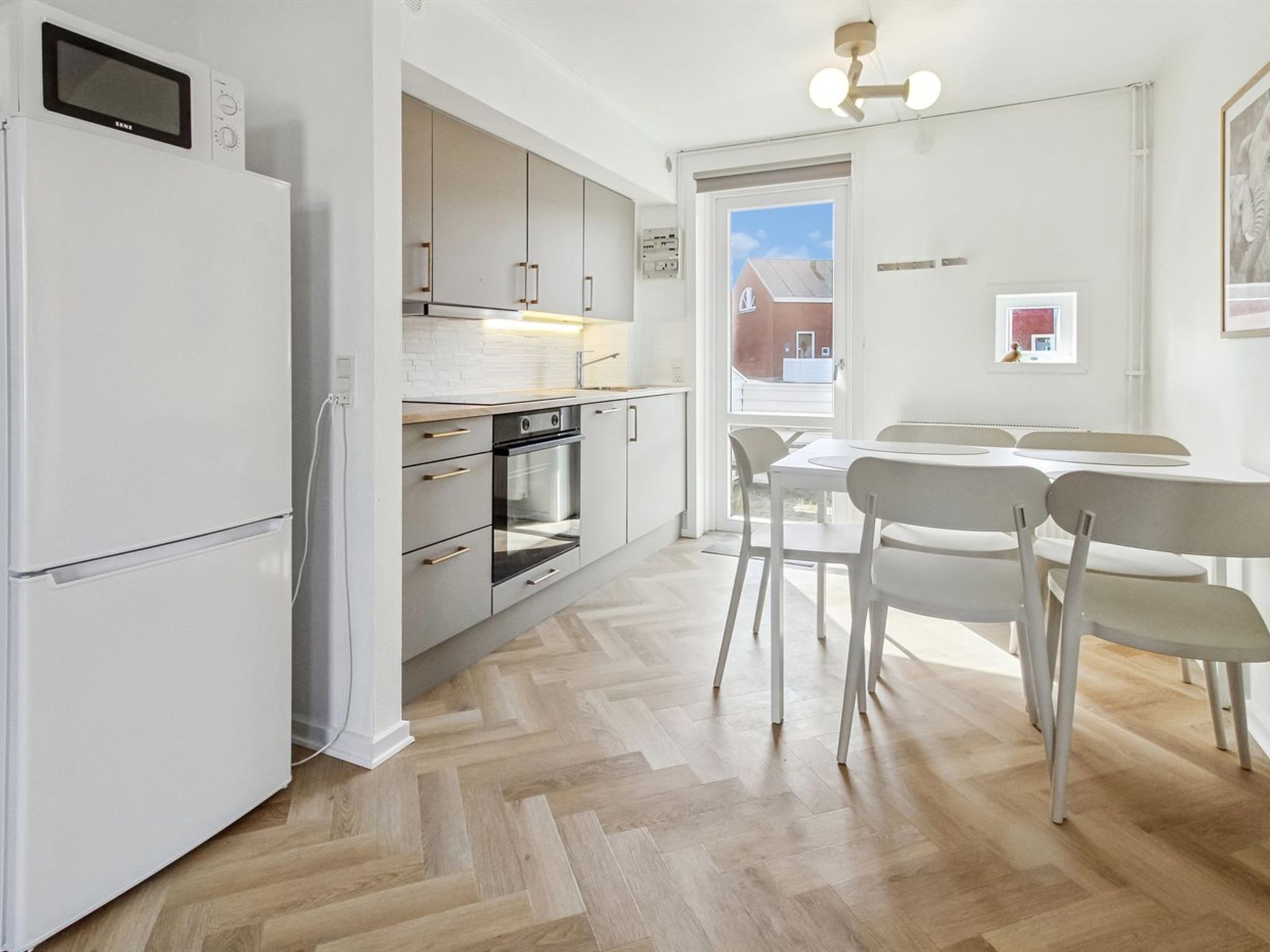 A modern kitchen with a refrigerator, oven, and microwave, a dining area with chairs and a table, and a view of the outside through a door.