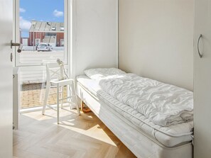 A small, single-bed room with a wooden floor, a white chair, and a window overlooking a street.