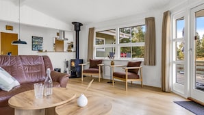 En hyggelig stue med pejs, lædersofa, sofabord i træ og store vinduer med udsigt til naturen.