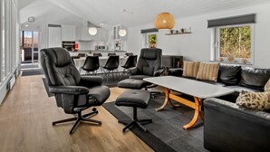 Moderni olohuone, jossa on suuri pöytä, mustat nahkaiset nojatuolit ja sohva. Taustalla näkyy keittiö, jossa on valkoiset kaapit, sekä ruokailutila, jossa on mustat tuolit.