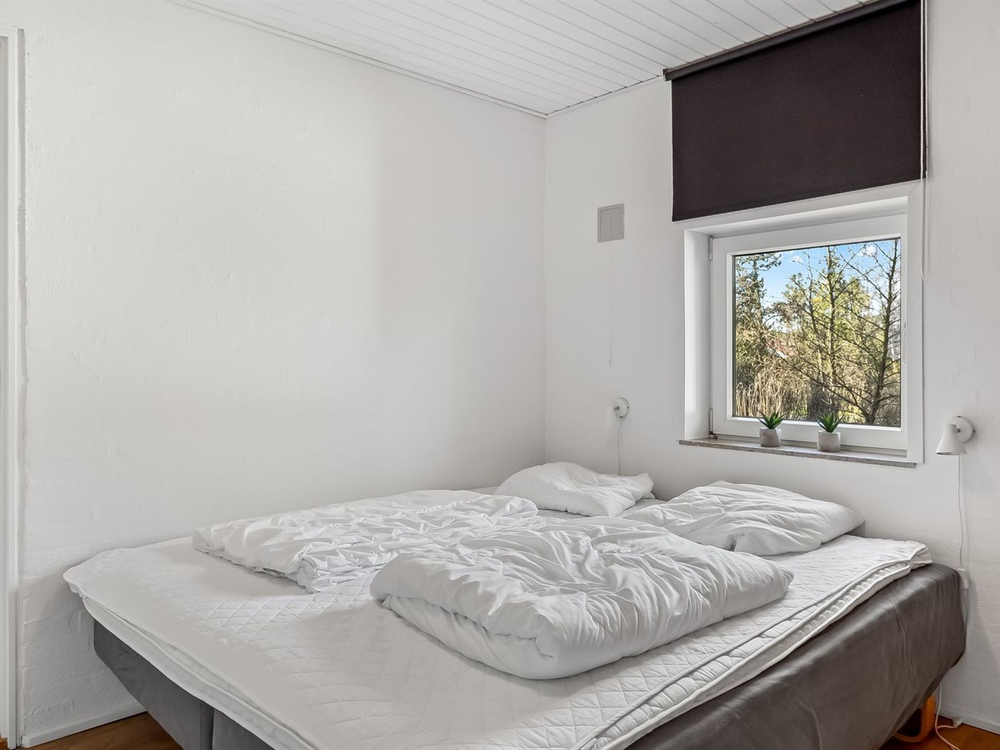 Een enkel bed met wit beddengoed in een kamer met witte wanden en een raam met uitzicht op bomen.