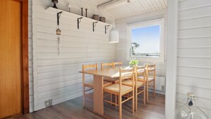 Een eetruimte met een houten tafel en stoelen, witte houten wanden, een raam met uitzicht en een plank met decoratieve objecten.