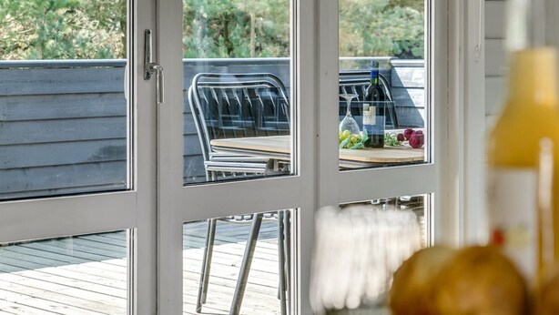 Uitzicht door een glazen deur op een terras met een tafel, stoelen en een fles wijn.