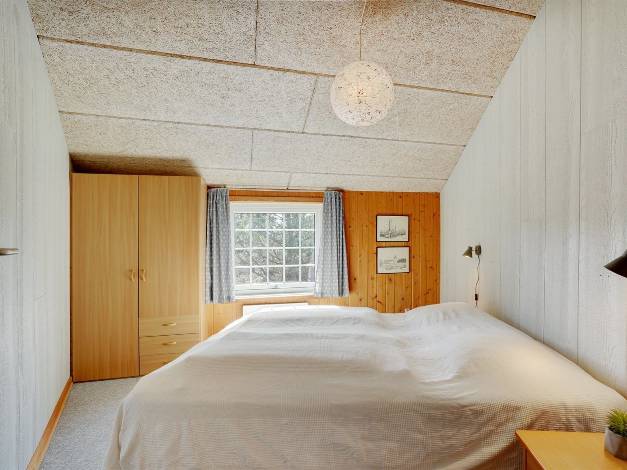 A bedroom with a large bed, wooden wardrobe, a window with curtains, and a ceiling light fixture.