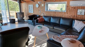 Ein gemütliches Wohnzimmer mit einer schwarzen Ledersofa, einem weißen Couchtisch und einem schwarzen Ledersessel. Ein Kaminofen, ein Fenster mit Blick auf Grünflächen und eine Wand-Klimaanlage sind ebenfalls vorhanden.