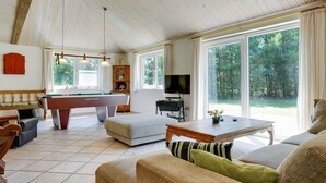 Ein geräumiges Wohnzimmer mit Billardtisch, bequemen Sitzgelegenheiten, Fernseher und großen Fenstern mit Blick ins Freie.