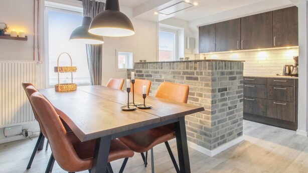 Ein moderner Essbereich mit Holztisch und Ledersesseln, einer Ziegelwand als Blickfang und dunklen Holzausziehtischen in der angrenzenden Küche.