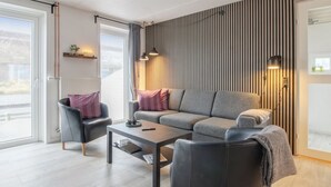 Ein modernes Wohnzimmer mit einem grauen Sofa, schwarzen Sesseln und einem Couchtisch. Es gibt zwei Pendelleuchten, ein Fenster mit Blick und eine Wand aus Holzlatten.
