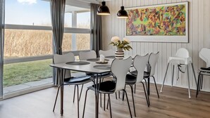 Un coin repas avec une table dressée pour quatre personnes, entourée de chaises blanches aux pieds noirs. On y trouve une grande fenêtre avec des rideaux, un tableau au mur et un vase rempli de fleurs sur la table.