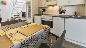 Une cuisine compacte avec des meubles blancs, une cuisinière et un évier. On y trouve également un coin repas avec une table et des chaises, ainsi qu’un escalier menant à une autre pièce.