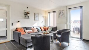 Een moderne woonkamer met een grijze bank, zwarte fauteuils en een salontafel. Er zijn twee ramen met zonwering, een deur en uitzicht op het buitenland.