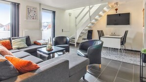 Een moderne woonkamer met een bank, stoelen, een salontafel en een eethoek met een tafel en stoelen. Een trap is op de achtergrond zichtbaar.