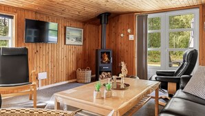 L’intérieur chaleureux d’un chalet avec un plafond en bois, une cheminée, un téléviseur fixé au mur et une grande fenêtre donnant sur le paysage extérieur.