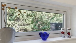 Une fenêtre donnant sur des arbres, un vase contenant des branches séchées, un vase en verre bleu et deux récipients en verre contenant des objets rouges.