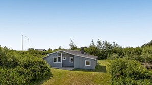 En etagers grå bygning med saddeltag, omgivet af frodig vegetation.