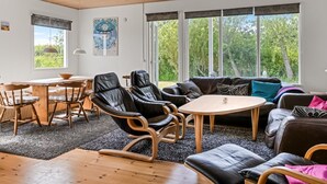 En moderne stue med træloft, spiseområde med et træbord og stole, en sort lædersofa og store vinduer med udsigt over grønne omgivelser.