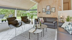Ein modernes Wohnzimmer mit einem grauen Sofa, zwei schwarzen Sesseln, einem Couchtisch und einem großen Fenster mit Blick auf Grünflächen.