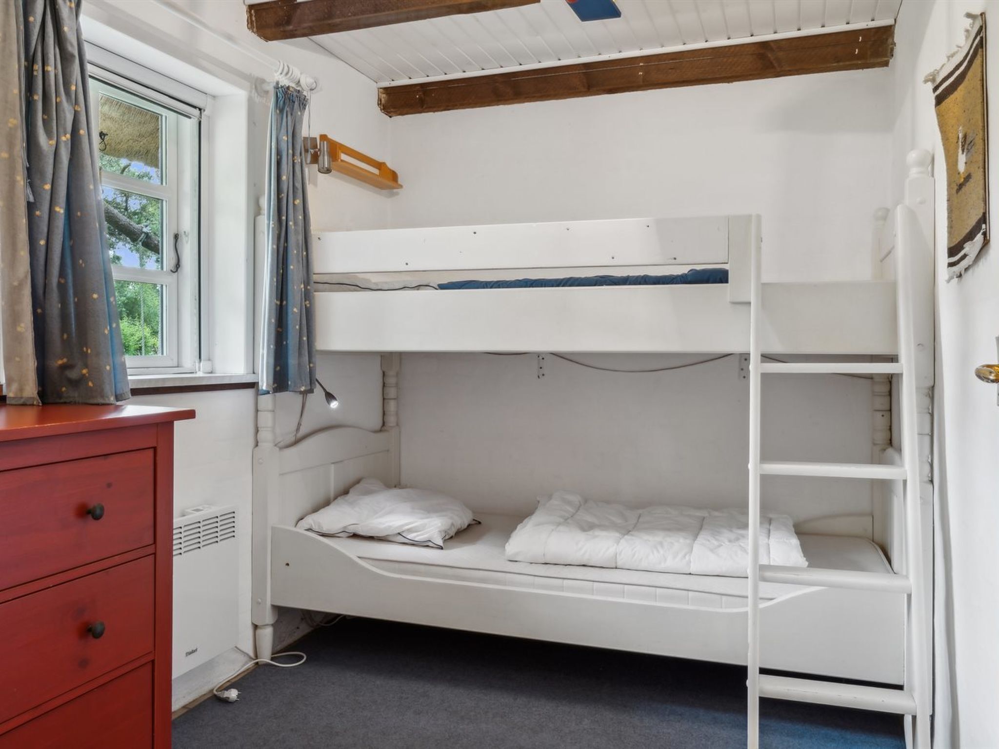 A bunk bed room with a red dresser, a window with curtains, and a wooden ceiling beam.