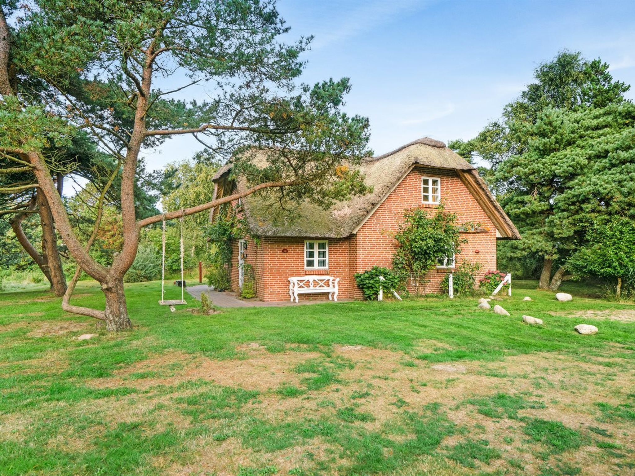 A brick house with a thatched roof, surrounded by trees and a grassy lawn.