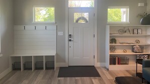 A home entryway with a white door, a bench with hooks, a small shelf with decorative items, and a window with a view of trees. - Modern, pet-friendly stay centrally located, minutes to everything in Bend! (Bend)
