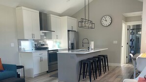 A modern kitchen with white cabinets, a central island with stools, a clock on the wall, and a dining area with a red table runner. - Modern, pet-friendly stay centrally located, minutes to everything in Bend! (Bend)