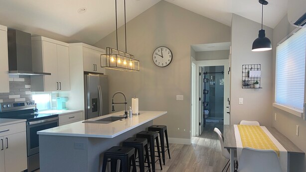 A modern kitchen with a central island, bar stools, a dining area with a table and chairs, a clock on the wall, and a window with blinds. - Modern, pet-friendly stay centrally located, minutes to everything in Bend! (Bend)